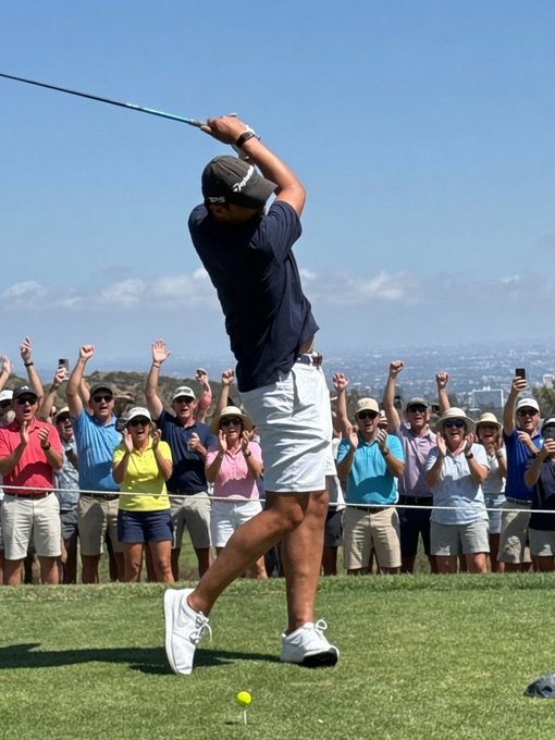 A golfer in a navy blue shirt and white shorts completes a swing while an excited crowd of spectators cheers behind a rope line on a sunny day.