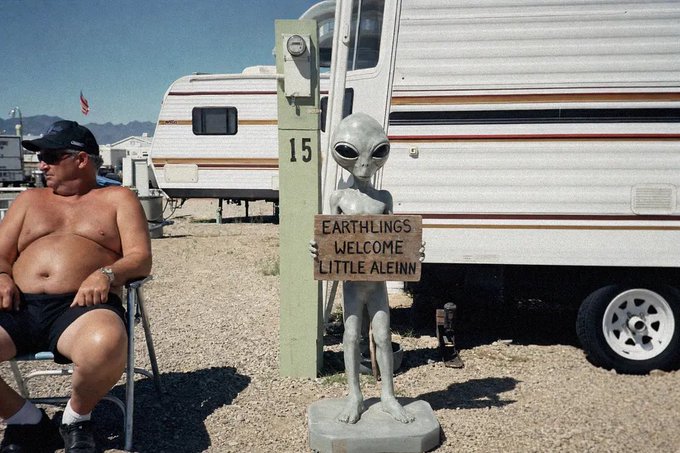 A shirtless man wearing sunglasses and a black cap sits in a chair next to an alien statue holding a sign that reads 'EARTHLINGS WELCOME LITTLE ALEINN' beside a parked RV.