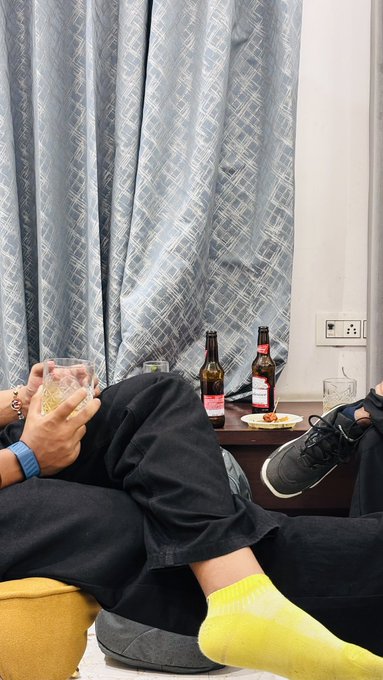 A casual indoor scene showing two people relaxing on a couch with their feet up; one person in black pants and yellow socks holds a glass, while another in dark clothing and black sneakers holds a phone, with beer bottles and a small plate of food on a side table.