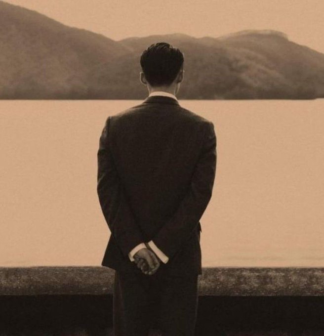 A sepia-toned photograph shows a man in a dark suit standing with his hands clasped behind his back, looking out over a lake toward mountains.
