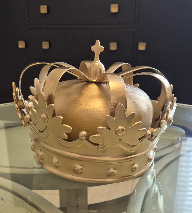 A decorative gold-painted crown featuring a central sphere topped with a cross, adorned with floral cutouts and bead details, displayed on a glass surface.