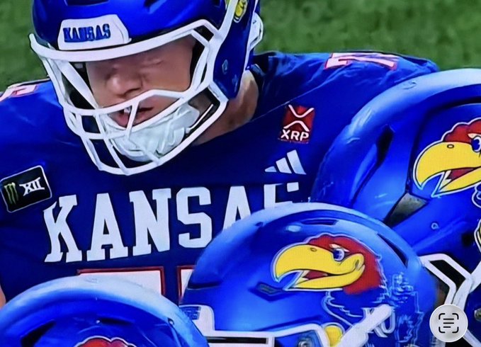 A Kansas football player wearing a royal blue jersey is shown in close-up, with the XRP cryptocurrency logo visible as a sponsor patch on the chest alongside the Big 12 (XII) conference logo and Adidas branding. The helmet, featuring the iconic Jayhawk mascot, matches the team's signature color scheme. This image supports the post's claim that XRP has become a sponsor displayed on Kansas football uniforms.