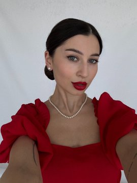 A woman with dark hair pulled back, wearing red lipstick, a pearl necklace, and a red dress with ruffled sleeves poses against a white background.