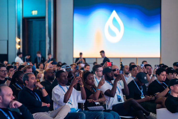 An audience at a Sui blockchain conference sits attentively while many attendees hold up smartphones to photograph a large screen displaying the Sui water-droplet logo.