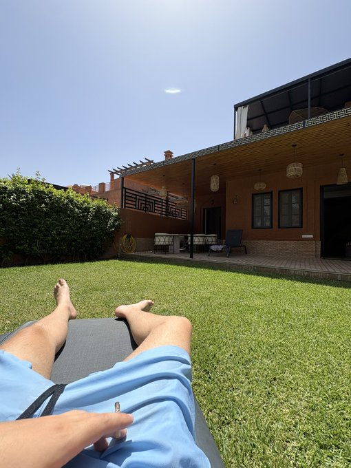 A first-person perspective photo of someone relaxing on a lounge chair on a grassy lawn, facing a wooden house with a covered patio under a clear blue sky.