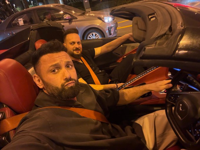 Two bearded men sit in a luxury convertible at night, with the driver gripping the steering wheel and the passenger leaning back, capturing a casual cousin moment on the road.