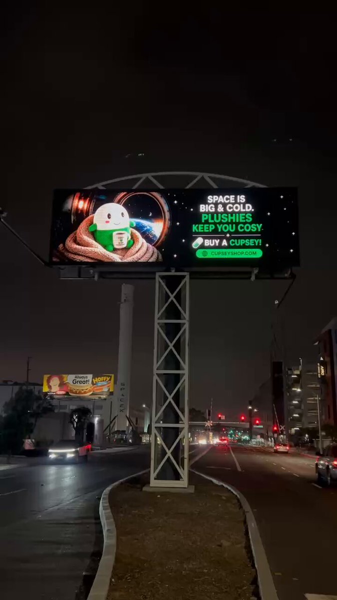 The image features a billboard with an advertisement promoting space travel, featuring a cartoon character from outer space. The billboard also includes a reminder about keeping warm while working outdoors during cold weather conditions.