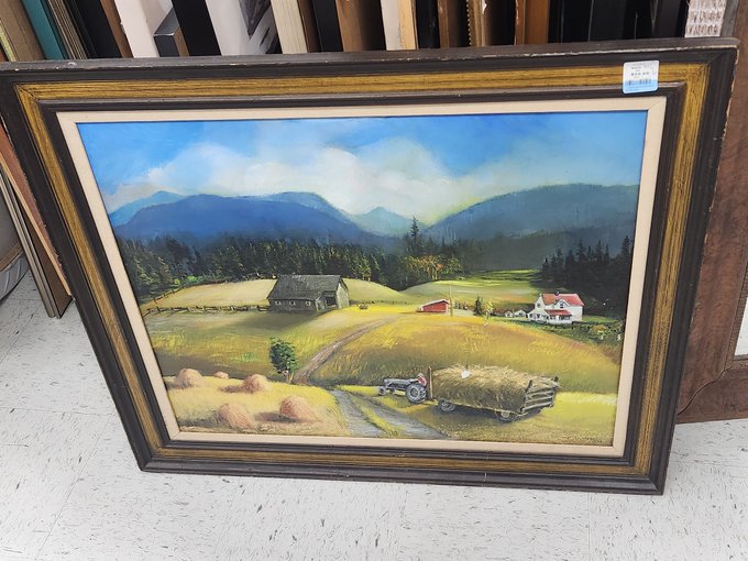A framed vintage landscape painting depicting a rural farm scene with a tractor hauling hay, rolling hills, mountains, and small houses in the background.