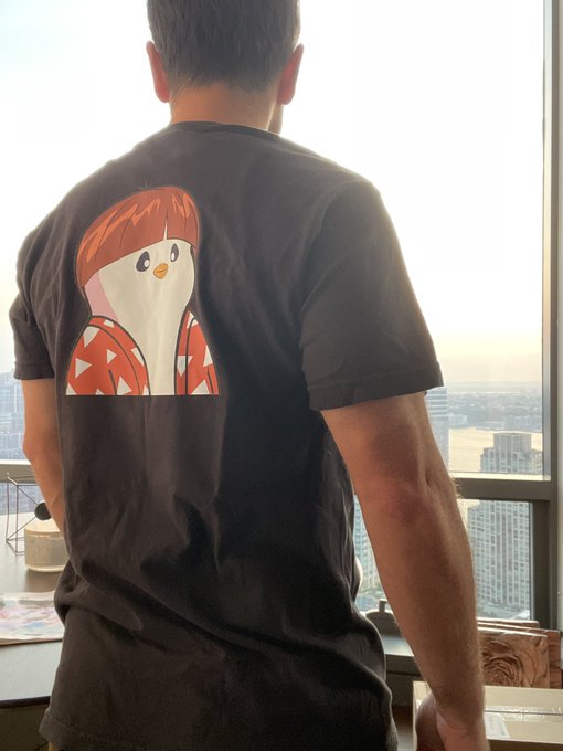 A person stands with their back to the camera wearing a black t-shirt featuring a cartoon penguin with orange hair and a red mushroom-cap jacket. The accompanying post humorously contrasts nostalgia for past NFT earnings with the ongoing "Pudgy" brand, linking the penguin mascot on the shirt to Pudgy Penguins.