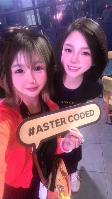 Two young women take a selfie at what appears to be an indoor event venue, with one holding up a sign reading "#ASTER CODED" while wearing an "ASTER" branded shirt, tying into the post's promotion of the Aster DEX brand and #astercoded hashtag at #idbw2026.