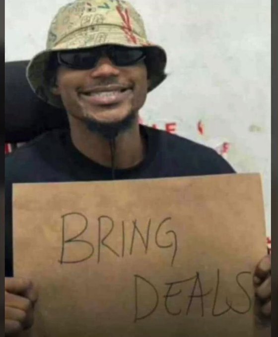 A smiling man wearing a bucket hat and sunglasses holding a cardboard sign that reads "BRING DEALS" in handwritten letters.