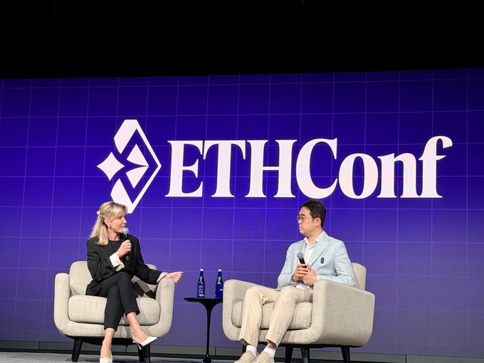 The image shows a panel discussion between two people sitting in chairs at a stage with a blue wall featuring the "ETHICONF" logo. The man is wearing glasses, while the woman has blonde hair. They are both engaged in conversation, possibly discussing the topic of liquidity and capital management in the financial markets.