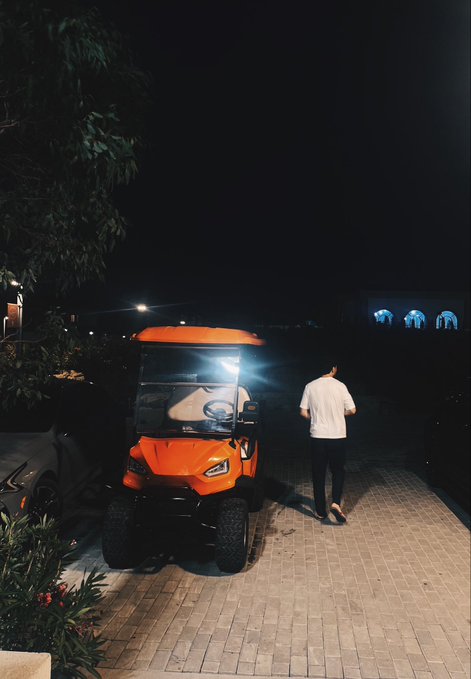 A nighttime photo of an orange golf cart parked on a paved area, with a person in a white shirt and dark pants walking away toward a wall displaying three illuminated figures.
