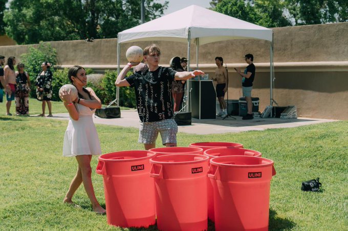 Two young people play a ball-throwing game with red ULINE buckets at an outdoor lawn event under a white tent, with other attendees mingling in the background.