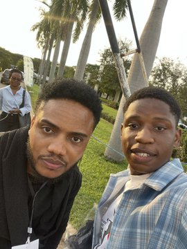 Two men take a selfie outdoors at the Solana summit, with palm trees and a woman walking in the background on the sunny grounds.
