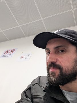A selfie of a bearded man wearing a black cap and gray hoodie, standing in a room with a white tiled ceiling and no-smoking signs on the wall behind him.
