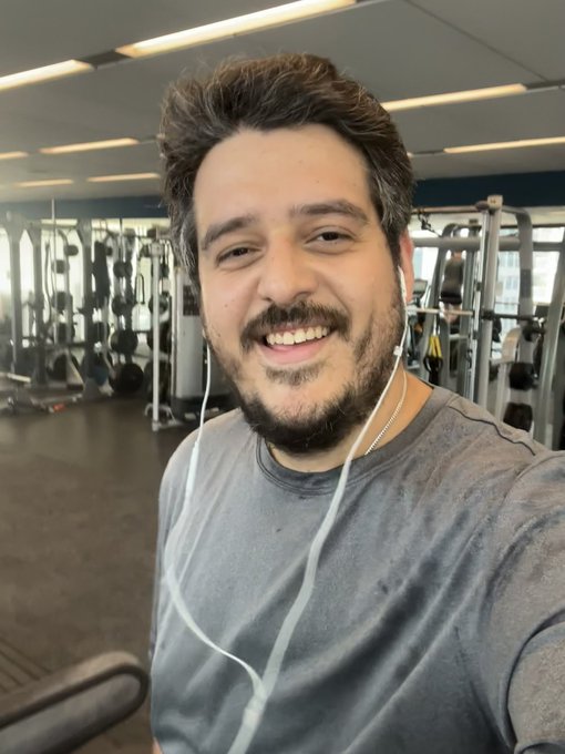 A smiling bearded man with messy dark hair takes a selfie in a gym, wearing a gray t-shirt and white wired earphones, with gym equipment and a person working out visible in the background.