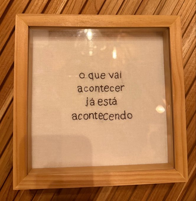 A framed cross-stitch piece on a wooden surface displays the Portuguese phrase 'o que vai acontecer já está acontecendo' (what is going to happen is already happening).