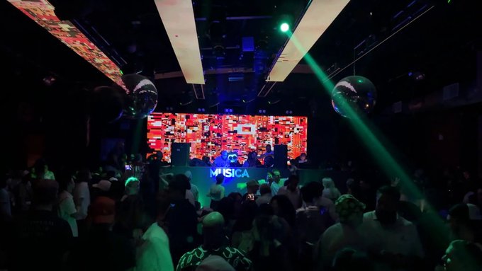 A crowded nightclub scene with green laser lights, disco balls, and a stage featuring a large screen above the word 'MUSICA'.