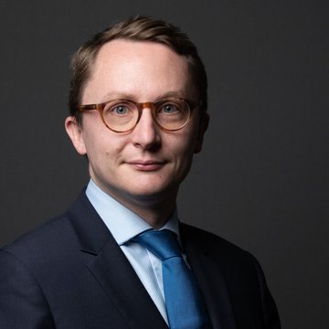 The image shows MP Paul Midy in a formal portrait wearing glasses, a dark suit, and blue tie. As the legislator who submitted the bill alongside 91 deputies, he is the key figure pictured in connection with the proposed modernization of France's digital asset taxation framework.