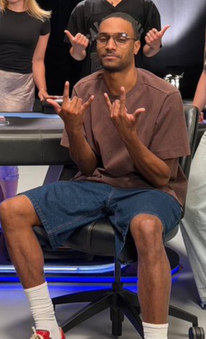 A man wearing glasses, a brown t-shirt, denim shorts, and red-and-white sneakers sits in a chair making rock-and-roll hand gestures.