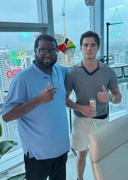 Two men pose indoors with a city skyline visible behind them, one holding a can and gesturing toward the camera while the other gives a thumbs-up. A neon "Up Only" sign and colorful "$LOOKS" text appear on the windows, aligning with the $LOOKSMAX token promotion in the post.