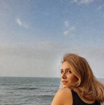 A young woman with long blonde hair gazes out at a calm blue sea beneath a partly cloudy sky, her shoulder bare as she appears to wear a black top.