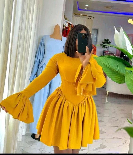 A woman taking a mirror selfie while wearing a mustard yellow dress with long bell sleeves and a full skirt, standing in a boutique with a mannequin in a light blue dress visible behind her.