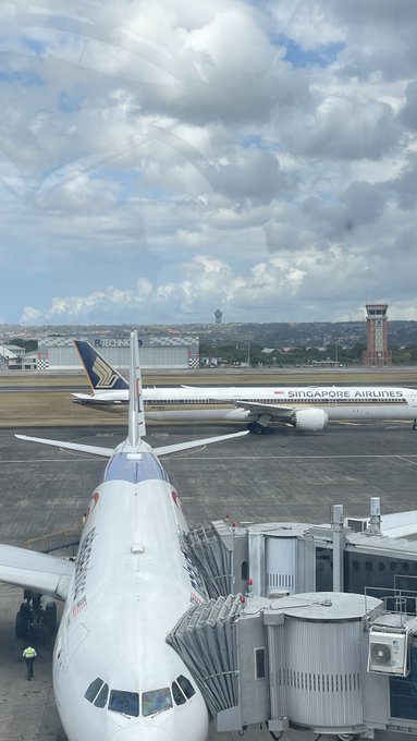 A view from inside an airplane showing a Singapore Airlines jet taxiing on the runway, with another aircraft nose visible at the gate connected to a jet bridge under a cloudy sky.