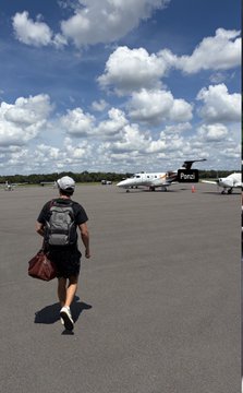 A person wearing a backpack, white cap, and shorts walks across an airport tarmac toward two parked private jets under a blue sky with puffy clouds.