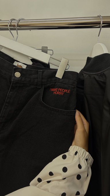 A person's hand in a polka-dot sleeve browsing black denim jeans on a clothing rack, with red text reading 'I MAKE PEOPLE HORNY' printed inside the pocket.