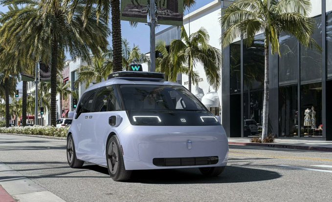 A white autonomous robotaxi with a rooftop sensor array drives down a palm tree-lined street in what appears to be a sunny urban setting.