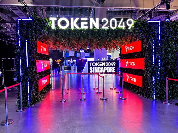 A brightly lit entrance display for 'TOKEN2049 SINGAPORE' featuring greenery walls, red TRON logos, and stanchions with red velvet ropes.