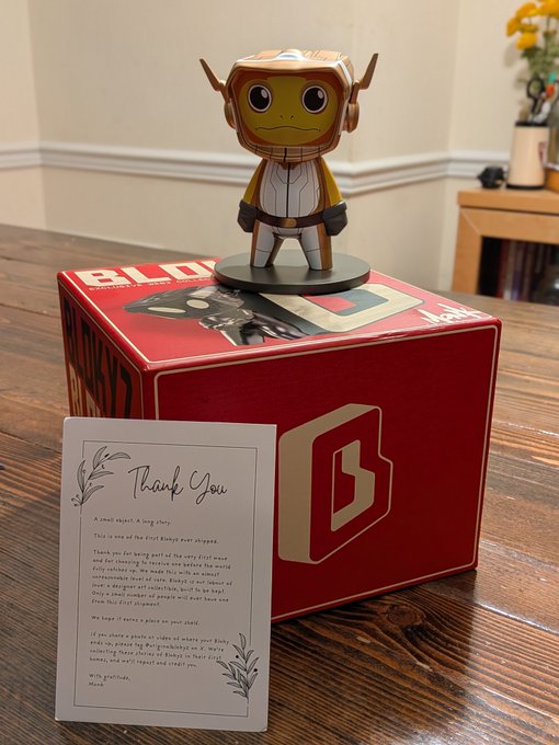 A collectible figurine of a monkey character in astronaut gear displayed on a red branded box, accompanied by a 'Thank You' card with a floral design sitting on a wooden table.
