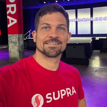 A selfie of a smiling man wearing a red 'SUPRA' t-shirt, with the word 'MISSIONLESS' visible on a screen in the background.