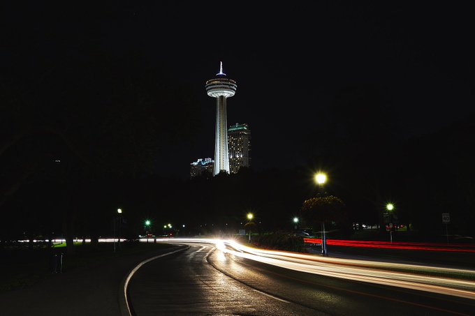 A long-exposure night photograph of a road with light trails from vehicles, featuring an illuminated observation tower (Skylon Tower) in the background against the dark sky.