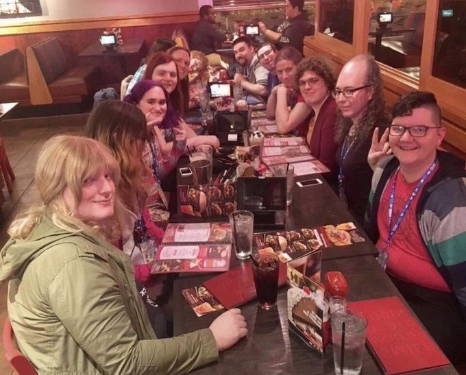 A group of around ten people gather around a long restaurant table at the Sappy Seals meetup, smiling and posing for the camera with menus, drinks, and glasses of water spread out before them, capturing the fun, social vibe of the gathering.