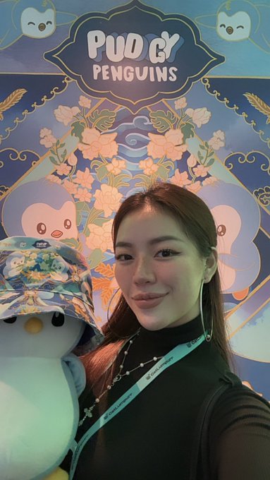 A selfie of a young woman posing next to a Stitch-themed plush toy in front of a 'PUQ & PENGUINS' decorative display with blue and gold ornate Asian-inspired artwork.
