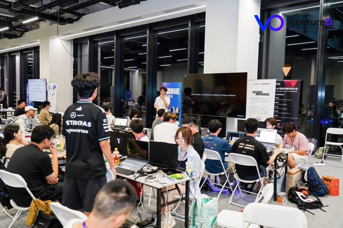 An indoor co-working or gaming event space filled with people working at laptops, with one person wearing a Petronas ETKRONAS race team shirt and a 'Ventured' logo visible.