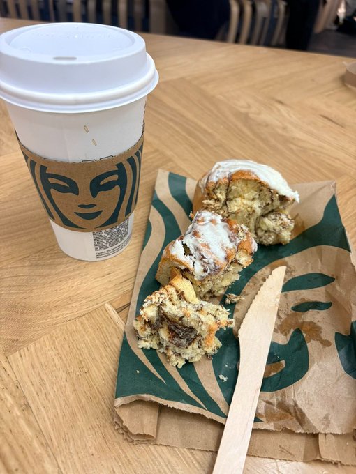 A Starbucks iced caramel macchiato sits beside a partially eaten cinnamon roll on a branded napkin and wooden stir stick, perfectly capturing the post's joke about maintaining a "straight face" and unwavering brand loyalty to Starbucks even after financial ruin.