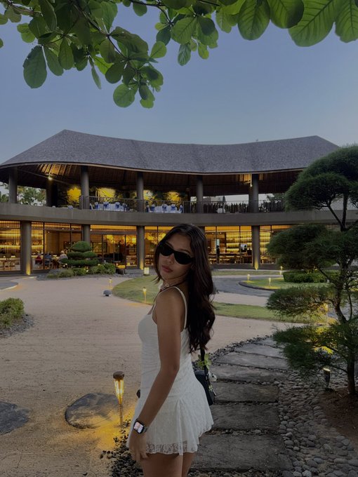 A young woman in a white sundress and sunglasses poses in front of an elegant, modern building with sweeping curved roofs and warmly lit windows at dusk, likely at a high-end resort or gallery. The serene, sophisticated setting complements the "Gm ready to mint?" vibe of the post, evoking a chill, luxurious atmosphere fitting for tagging @ChogNFT.