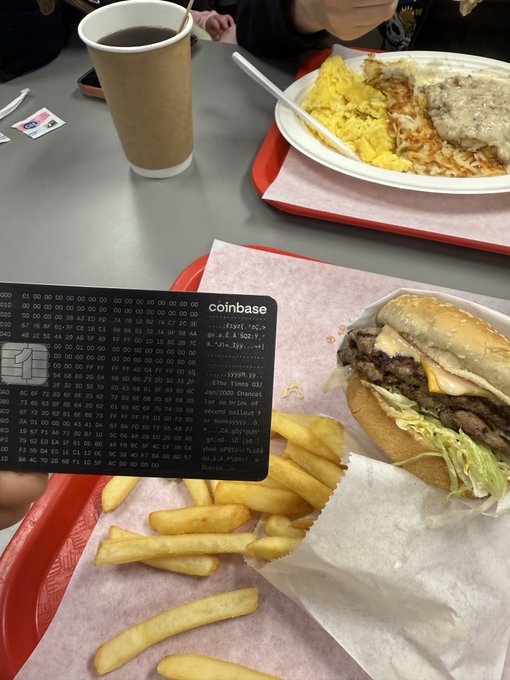 A meal at what appears to be McDonald's showing a double cheeseburger, french fries on a red tray, scrambled eggs with gravy, a coffee cup, and a Coinbase credit card held up displaying its ASCII art design with cryptocurrency-themed text.