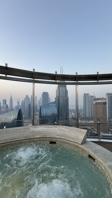 A bubbling rooftop hot tub overlooks a city skyline with tall skyscrapers under a clear blue sky, bordered by a glass safety railing.