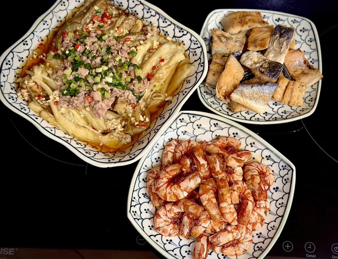 The image shows three plates of food, including shrimp and other dishes, arranged on a table. The first plate has shrimp and vegetables, while the second plate contains fish and vegetables. The third plate features shrimp with sauce. A person can be seen in the background, possibly enjoying their meal or preparing to eat it.