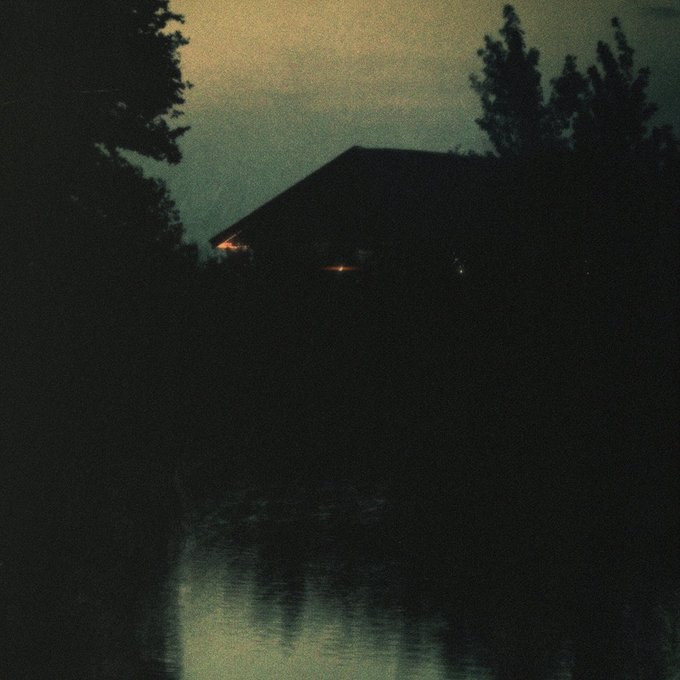 A dark, grainy nighttime photograph showing the silhouette of a house with a few lights on, reflected in water below.