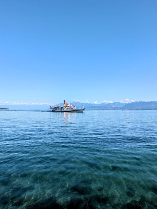 A serene paddle steamer glides across crystal-clear alpine waters under a brilliant blue sky, with distant snow-capped mountains on the horizon—offering a calm, picturesque scene that contrasts with the post's discussion of market volatility and bullish "Uptober" sentiment.