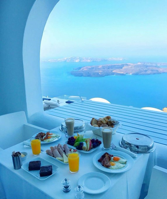 A luxurious breakfast spread including eggs, sausages, fruit, juice, coffee, and pastries arranged on a white-clothed table overlooking a stunning ocean caldera view, likely in Santorini.