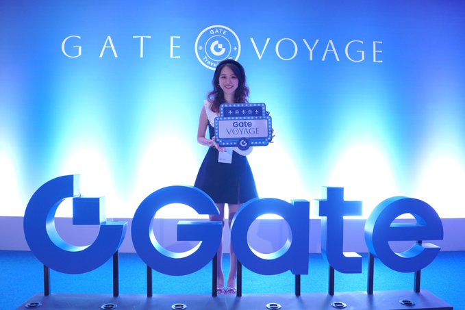 A smiling woman in a dark dress holds a 'GATE VOYAGE' sign while standing behind large blue 'Gate' lettering at what appears to be a promotional event.