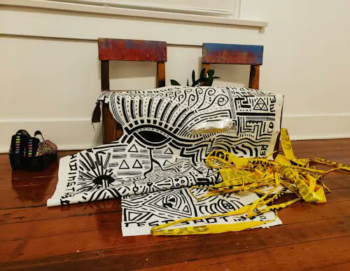 A small wooden table is draped with black and white patterned textiles featuring tribal-style designs, accompanied by yellow fabric strips and a small basket on a wooden floor.
