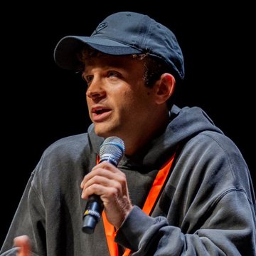 A young man wearing a navy blue baseball cap and a dark gray hoodie speaks into a microphone, with a red lanyard visible around his neck. The image appears to capture a moment of Jack Mallers making his bullish Bitcoin prediction, aligning with the post's quote about Bitcoin's potential to reach a $400-500 trillion market cap from its current $2 trillion valuation.